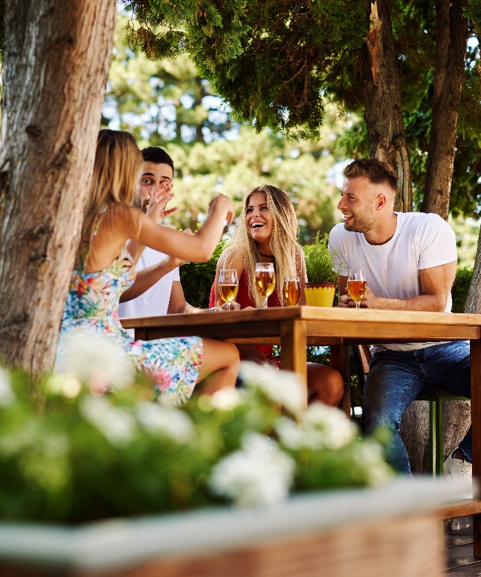 Group of young people sitting at a table in a summer bar laughing and drinking beer